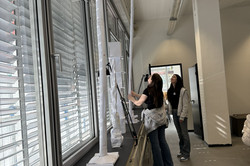 Two girls are standing in front of a windowsill, on the windowsill are towers that they have built out of paper.