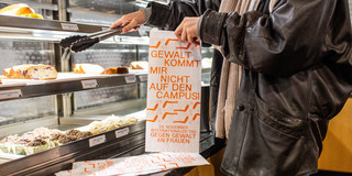 Eine Person bedient sich in der Cafeteria, die Brötchentüte hat die Aufschrift "Gewalt kommt mir nicht auf den Campus"