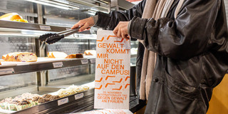 Eine Person bedient sich in der Cafeteria, die Brötchentüte hat die Aufschrift "Gewalt kommt mir nicht auf den Campus"