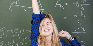 Mädchen an Tafel A girl stands in front of a blackboard with mathematical formulae. She raises one arm in the air. She is happy.