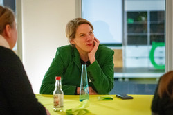 Person mit grünem Blazer sitzt an einem Tisch mit gelber Tischdecke und hält das Kinn mit der Hand gestützt.