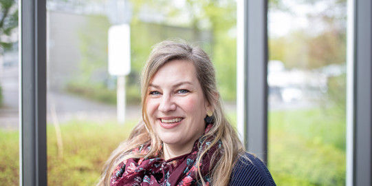 Lächelnde Frau mit langen blonden Haaren, dunkelblauem Pullover und gemustertem Schal vor Glasfenstern mit grünem Hintergrund.