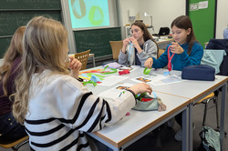 Aus bunten Pappstreifen basteln vier Schülerinnen ein eigenes Möbiusband. Sie sitzen um einen Tisch, im Hintergrund ist das Möbiusband auf einer Leinwand abgebildet.
