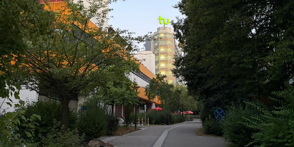Ein gepflasterter Weg führt zwischen dichtem Grün und Gebäuden entlang. Im Hintergrund ragt ein mehrstöckiges Gebäude mit einer auffälligen, leuchtend grünen Skulptur auf dem Dach in den Himmel. Die Szene ist von Bäumen und Sträuchern gesäumt, der Himmel ist klar und hellblau.