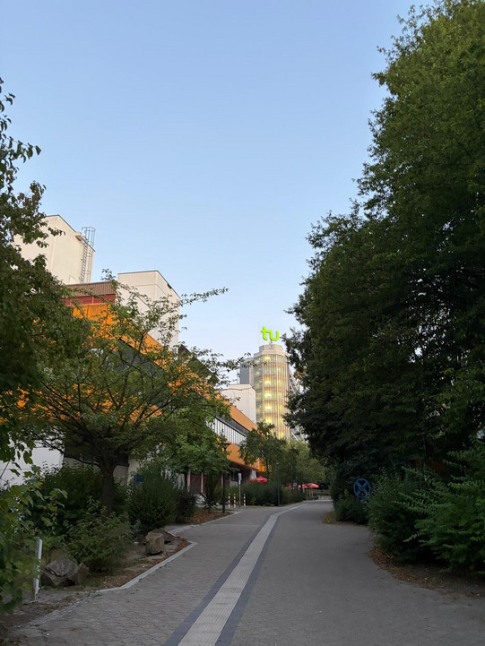 Ein gepflasterter Weg führt zwischen dichtem Grün und Gebäuden entlang. Im Hintergrund ragt ein mehrstöckiges Gebäude mit einer auffälligen, leuchtend grünen Skulptur auf dem Dach in den Himmel. Die Szene ist von Bäumen und Sträuchern gesäumt, der Himmel ist klar und hellblau.