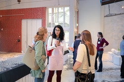 Drei Frauen stehen in einem hellen Raum und unterhalten sich, im Hintergrund zwei weitere Personen und ein Tisch mit vielen Weingläsern.