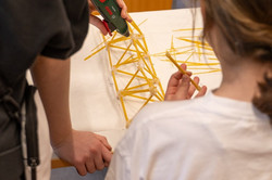 Zwei Personen bauen mit Spaghetti und Heißklebepistole eine filigrane Brückenkonstruktion auf einem Tisch.