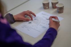 Photographed from above: A girl is sitting in front of a worksheet. There is a cube on the sheet and she is holding a pencil in her right hand. 