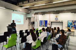 Ten schoolgirls sit on chairs in five rows of chairs. The workshop leader stands in front of them. An image is projected onto a screen in the background. It says ‘Welcome. Biological and chemical engineering’. 