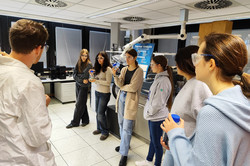 Six schoolgirls stand in a row. They are wearing safety goggles. The workshop leader is on the left of the picture.