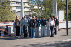 Gruppe von jungen Menschen mit Rucksäcken steht auf einem Gehweg im Freien vor einem Geländer.