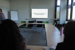 Konferenzraum mit U-förmigem Tisch, mehreren Personen und Bildschirm mit Text 'Wie werde ich Lehrer an der Berufsschule'.