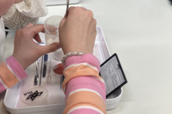 Junges Mädchen mit gestreiftem Pullover arbeitet an einem Experiment mit einem Plastikbecher und einem Löffel auf einem weißen Tablett mit DLR-Logo.