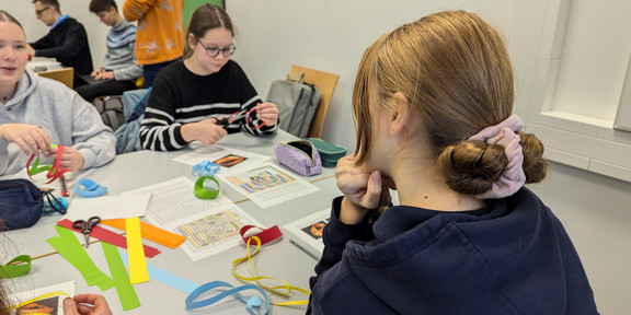 Three girls are sitting around a table. There are lots of worksheets and strips of paper in four different colours on the table. 