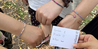 Mehrere Hände mit bunten Armbändern bilden einen Kreis, eine Hand hält eine Karte mit der Aufschrift 'Grün 1 Pink 0 blau - excellent IT'.