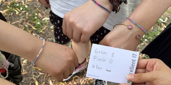 Mehrere Hände mit bunten Armbändern bilden einen Kreis, eine Hand hält eine Karte mit der Aufschrift 'Grün 1 Pink 0 blau - excellent IT'.