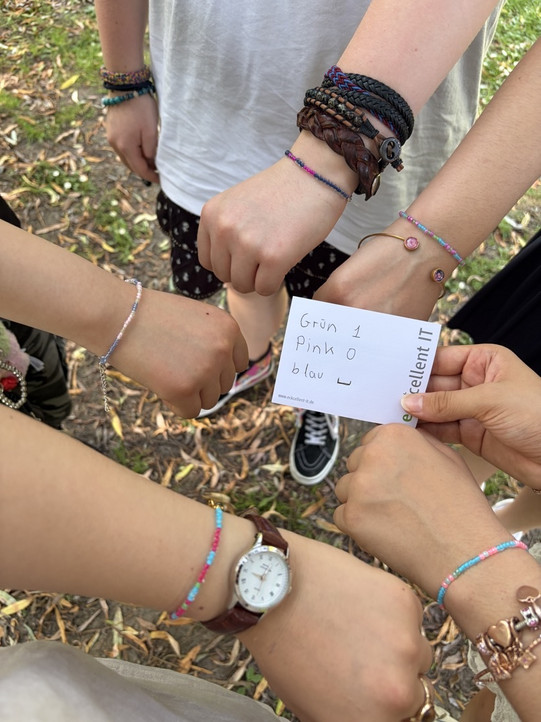 Mehrere Hände mit bunten Armbändern bilden einen Kreis, eine Hand hält eine Karte mit der Aufschrift 'Grün 1 Pink 0 blau - excellent IT'.