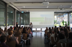 Vortragssituation in einem hellen Raum mit Publikum, zwei Personen sprechen vor einer Leinwand mit Präsentation.