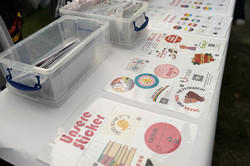 Tisch mit durchsichtigen Boxen und bedruckten Aufklebern, im Hintergrund Personen bei einem Sommerfest.
