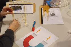 On a table lies a sheet with a magnet on it. Girls stick spaghetti into marshmallows.
