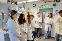 Six schoolgirls stand in a row. They are wearing white coats and protective goggles. 