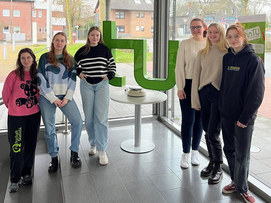 Die sechs Mentorinnen stehen zusammen für ein Gruppenfoto. In der Mitte steht ein grünes TU Logo auf einem Tisch. 