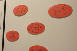 Fünf orangefarbene ovale Zettel mit einfachen schwarzen Strichzeichnungen an einer weißen Wand.