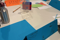 Mehrere aufgestellte Namenskarten aus farbigem Papier auf einem Tisch, eine blaue Karte mit handschriftlichen Notizen und kleinen Zeichnungen im Vordergrund.