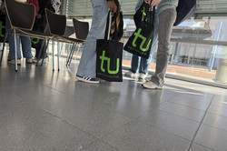 Photographed from below:Three girls are standing together. They are holding black jute bags with the green TU logo on them. 