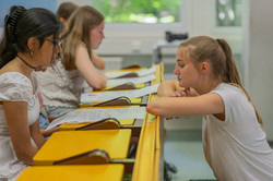 Ein Mädchen sitzt in einem Hörsaal. Vor ihr liegt eine Zeitung. Eine junge Frau kniet vor dem Tisch und erzählt etwas. 