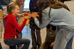 Five girls hold their hands together in a circle and balance one spaghetti noodle on them. 