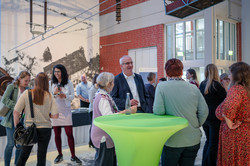 Mehrere Menschen stehen und unterhalten sich an einem runden Stehtisch in einer Halle mit Backsteinwand und großem Wandbild im Hintergrund.