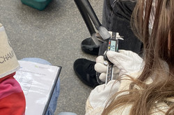 Junge Person mit langen Haaren und weißem Handschuh misst mit einer digitalen Schieblehre an einem Metallobjekt, daneben eine Person mit rotem Pullover und Papierhut mit der Aufschrift 'CHAMP'.