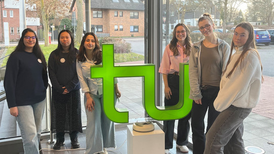 Six female students stand in a modern building with large windows behind a green stylized 'tu' logo on a white pedestal.