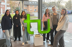 Sechs Studentinnen stehen in einem modernen Gebäude mit großen Fenstern hinter einem grünen, stilisierten 'tu'-Logo auf einem weißen Sockel.
