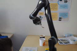Roboterarm mit Greifwerkzeug über einem Tisch mit bunten Bausteinen und einem Blatt Papier, eine Person steuert den Arm mit einem Handgerät.