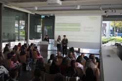 Vortragssituation in einem modernen Glasgebäude mit Publikum und drei Personen vor einer Leinwand mit Präsentationstext.