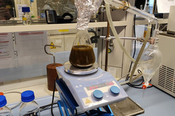 A flask is standing on a small lifting platform in a chemistry laboratory. The flask contains a brown liquid. The top of the flask is wrapped in aluminium foil and a glass tube leads from the foil into a glass funnel.