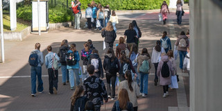 Gruppe von Menschen mit Rucksäcken und Taschen, die auf einem gepflasterten Weg zwischen Bäumen und Gebäuden gehen.