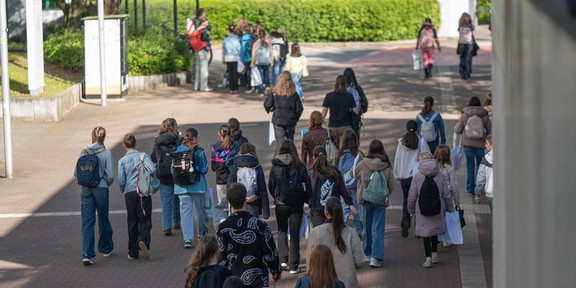 Gruppe von Menschen mit Rucksäcken und Taschen, die auf einem gepflasterten Weg zwischen Bäumen und Gebäuden gehen.