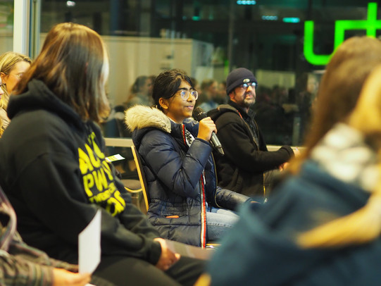 Ein Mädchen sitzt auf einem Stuhl und spricht in ein Mikrofon, welches sie in der rechten Hand hält. Rechts und links von ihr sind am Rand verschwommen andere Personen aus dem Publikum zu erkennen.