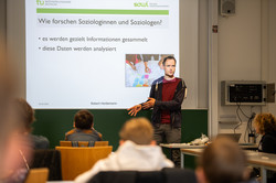 A man stands in front of a screen and talks. A presentation shows the question ‘How do sociologists do research?’