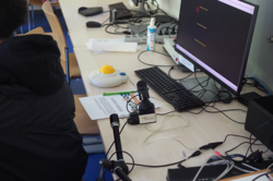 Arbeitsplatz mit Computer, Tastatur, Joystick und weiteren technischen Geräten an einem langen Tisch in einem Raum mit Stühlen und Schränken.