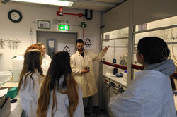 The workshop leader points with his left hand to the devices and the formulae behind or on the glass pane. Five girls look at the disc. 