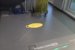 Nahaufnahme eines 3D-Druckers, der ein gelbes Objekt mit einer Öse auf einer Druckplatte herstellt.