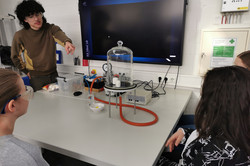 Three girls are sitting around a table. The workshop leader explains the experimental setup to the girls.