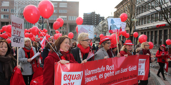 Women dressed in red draw attention to unequal pay with balloons and banners