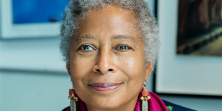 Portrait von Alice Walker: eine Schwarze Frau mit kurzen, grauen Haaren. Sie trägt eine dunkle Bluse mit lilanem Halstuch und hält eine Orangene Rose in der Hand.