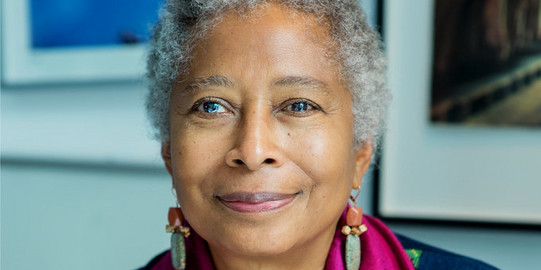 Portrait of Alice Walker: a black woman with short grey hair.She wears a dark blouse with a purple scarf and holds an orange rose in her hand.