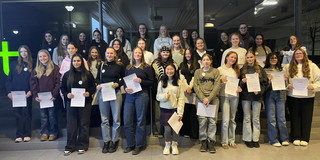 Eine große Gruppe von Mädchen und jungen Frauen steht in mehreren Reihen in einem Innenraum. Die meisten halten Papiere (Zertifikate) in den Händen. Die Kleidung ist vielfältig, darunter helle und dunkle Oberteile sowie Jeans. Im Hintergrund sind große Fenster und eine Decke mit sichtbaren Rohren und Lampen zu sehen.